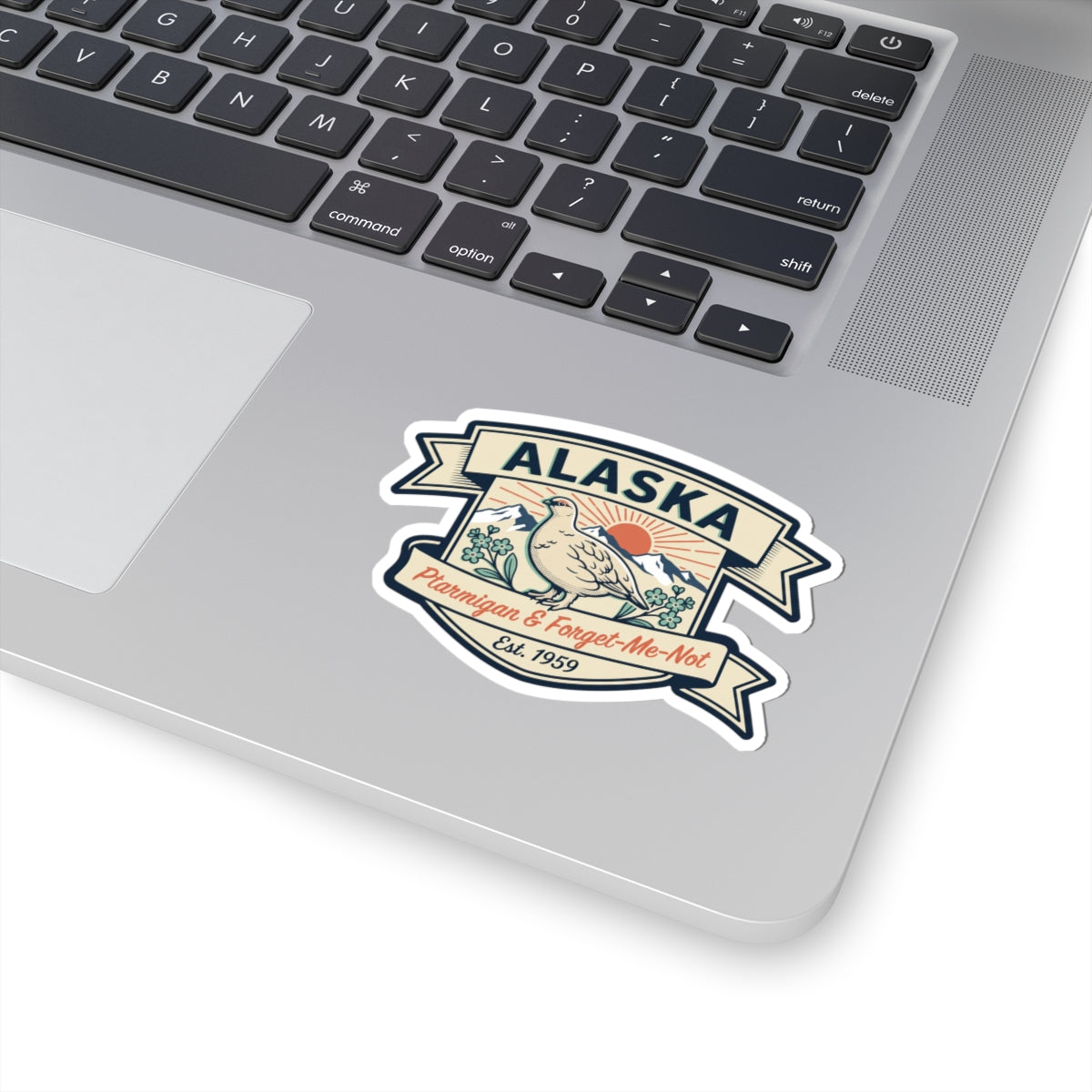Die-cut Alaska badge sticker on a gray laptop lid with ptarmigan and Est. 1959