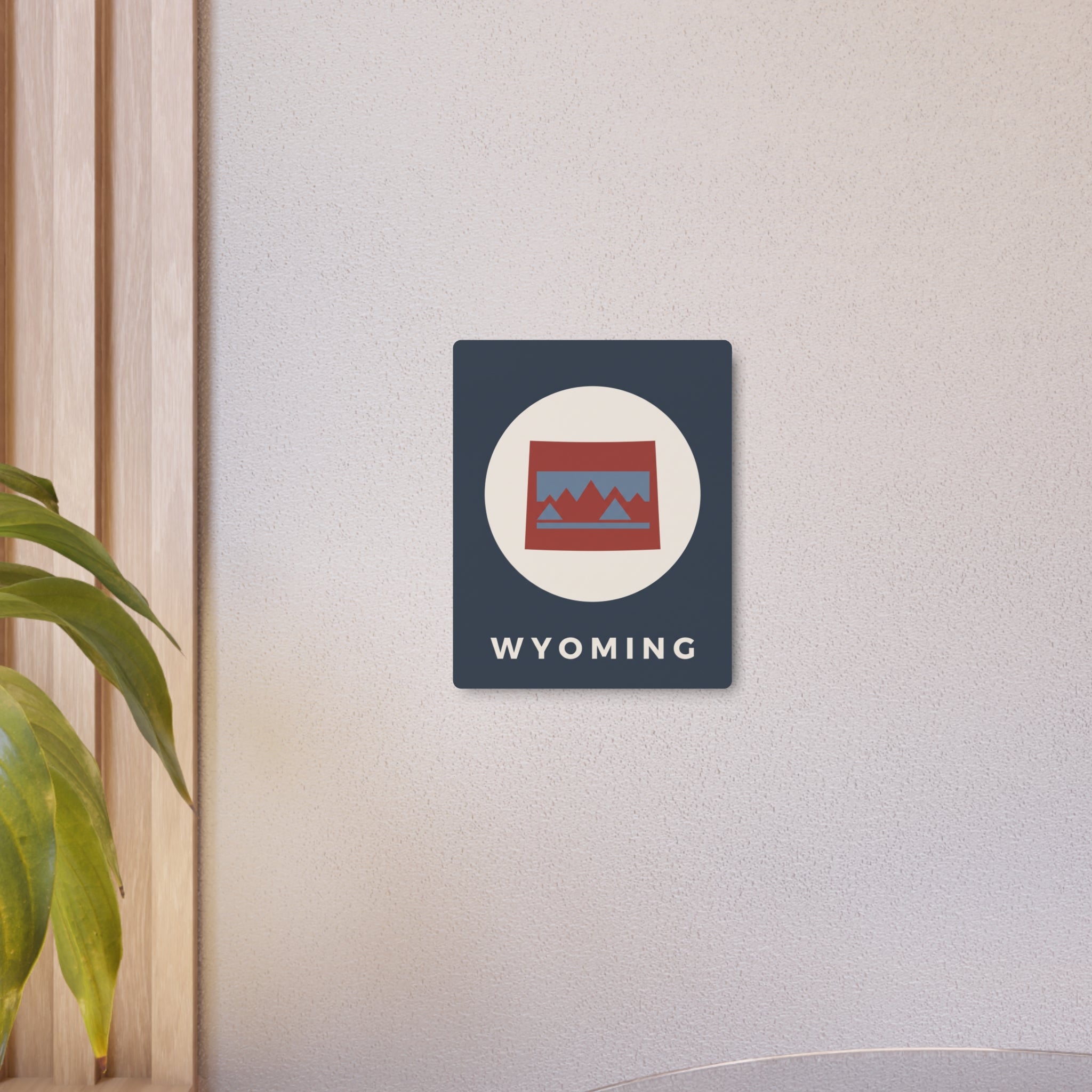 Wyoming state metal art sign on wall, dark blue square with oval mountains logo and “WYOMING”.